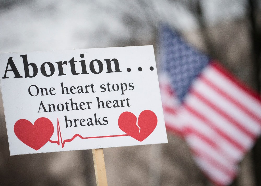 A rally sign reading "Abortion ... One heart stop, another heart breaks" with an American flag in the background