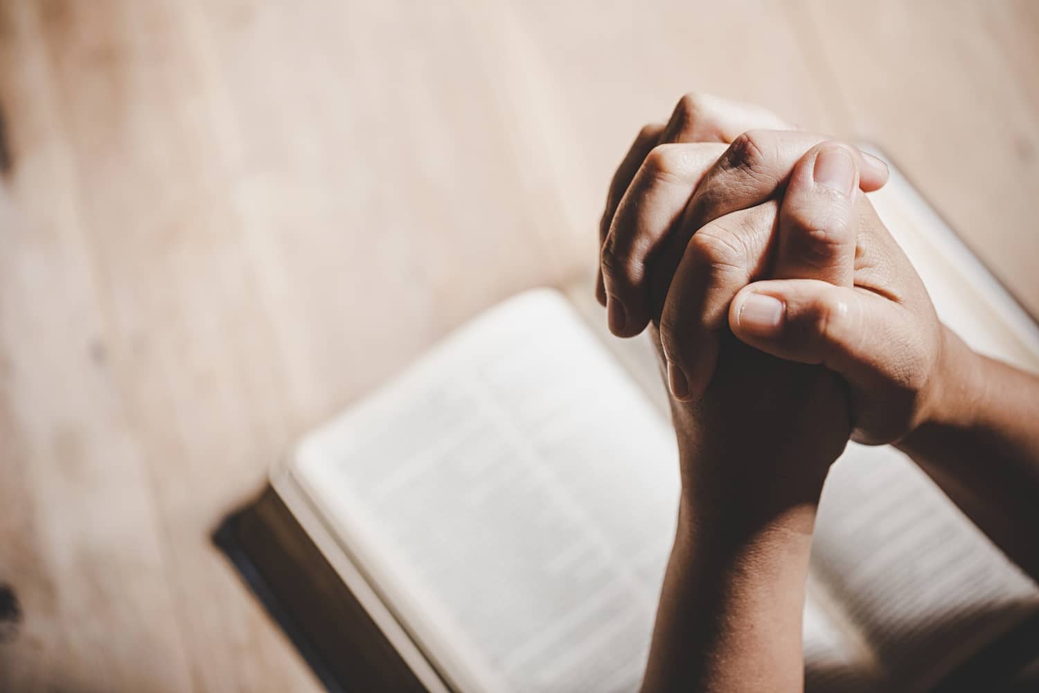 A photo of folded hands in prayer with a Bible in the background.